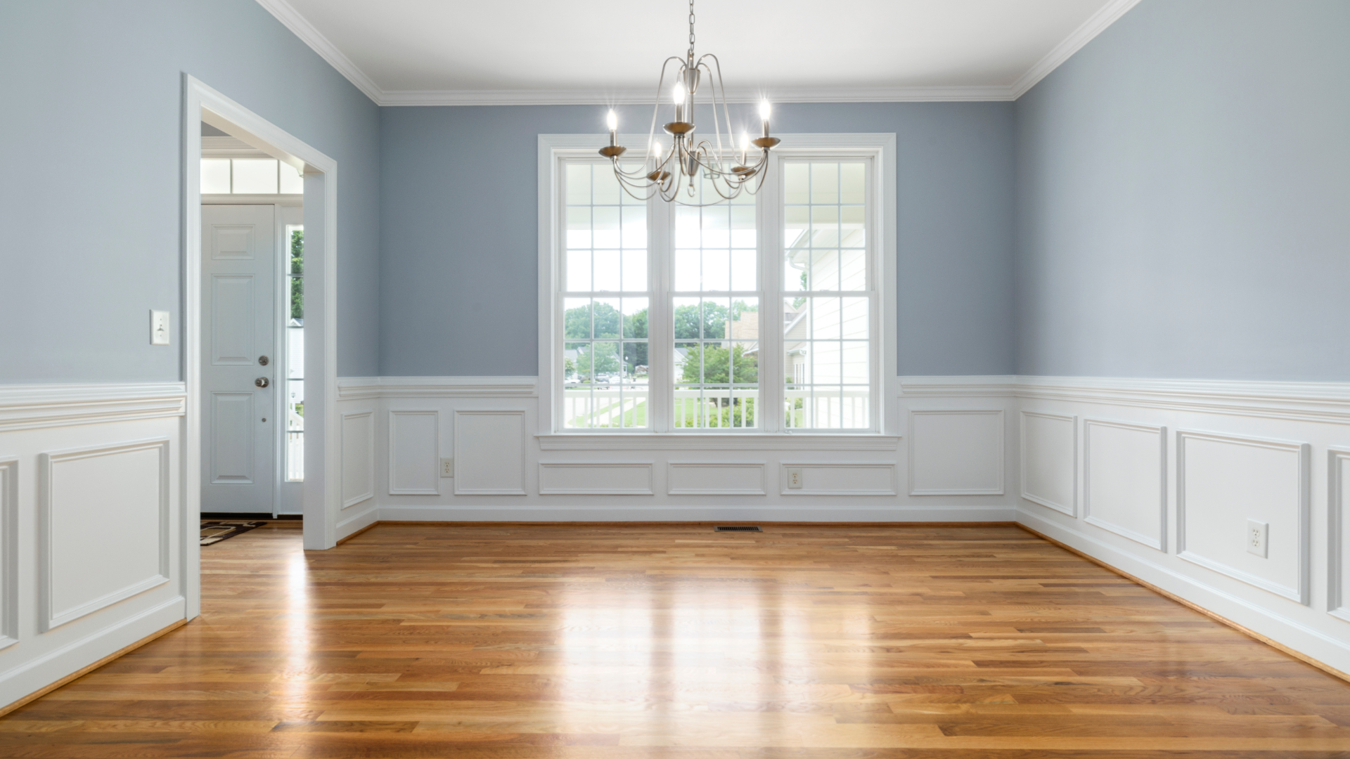 Empty dining room with light blue walls, white trim, and hardwood floors. A chandelier hangs above the window.