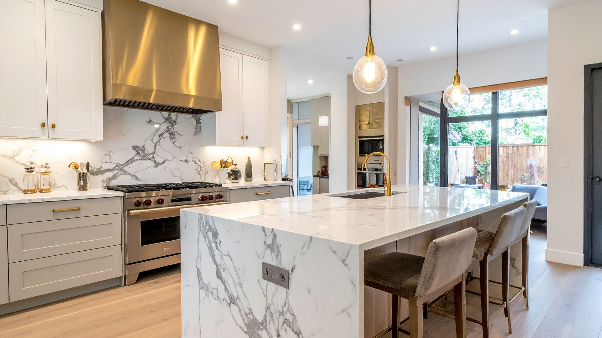 A modern kitchen with a marble island, gold accents, and light wood floors. Sunlight streams in from a patio door.