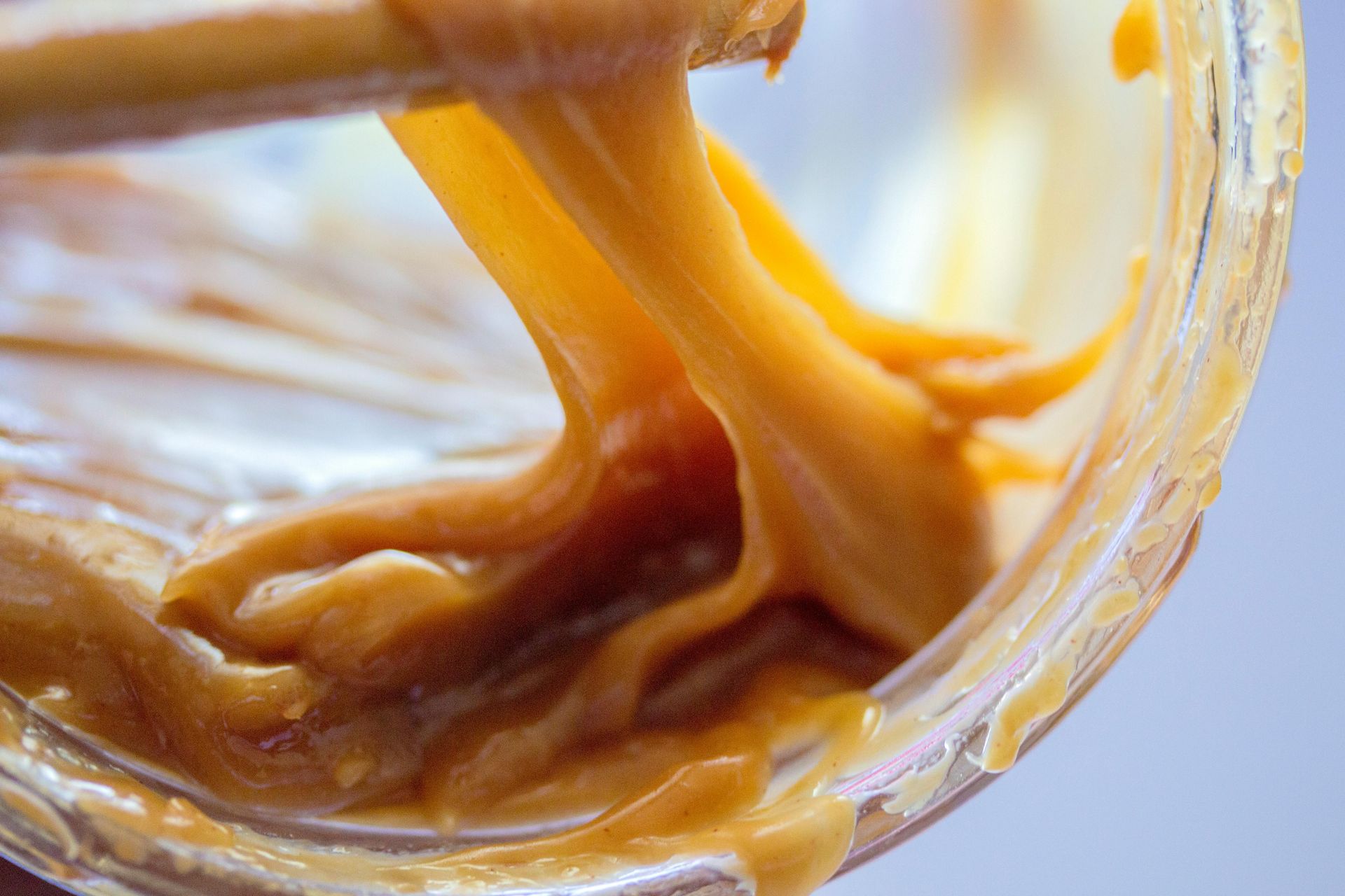 A wooden utensil pulls thick, stretchy golden-brown caramel from a clear glass jar.