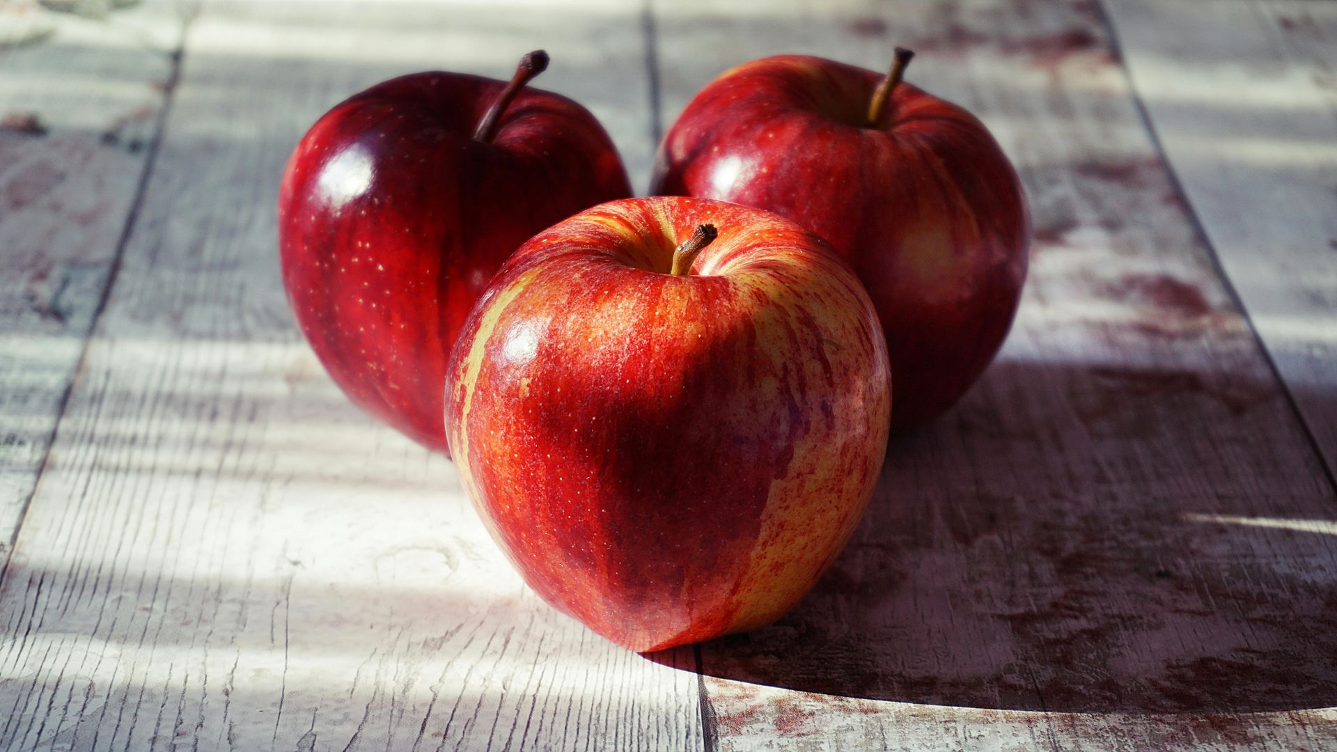 Three glossy red apples arranged in a triangle on a rustic, weathered wooden surface.