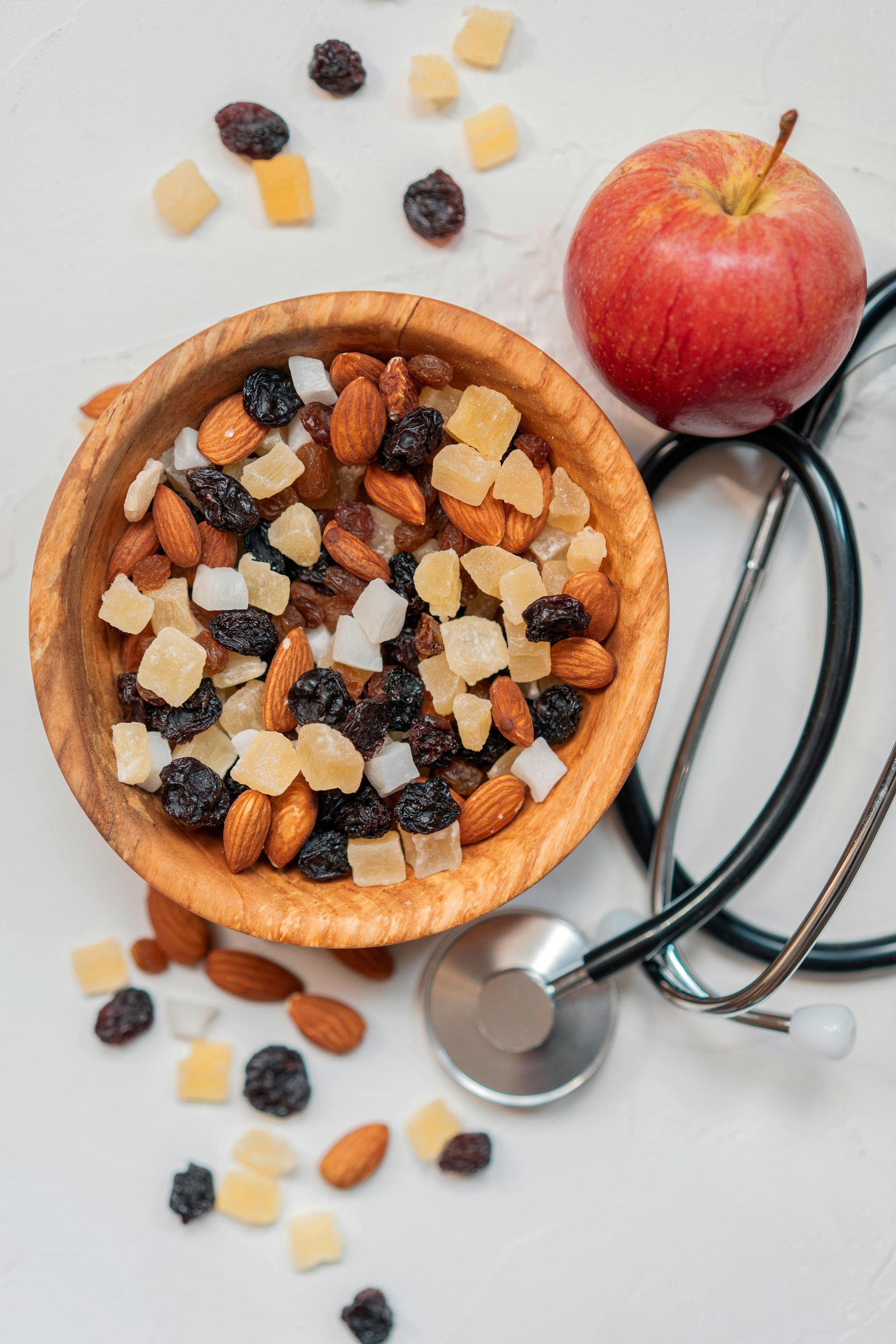 A wooden bowl of almonds and dried fruit, a fresh red apple, and a stethoscope on a light surface.