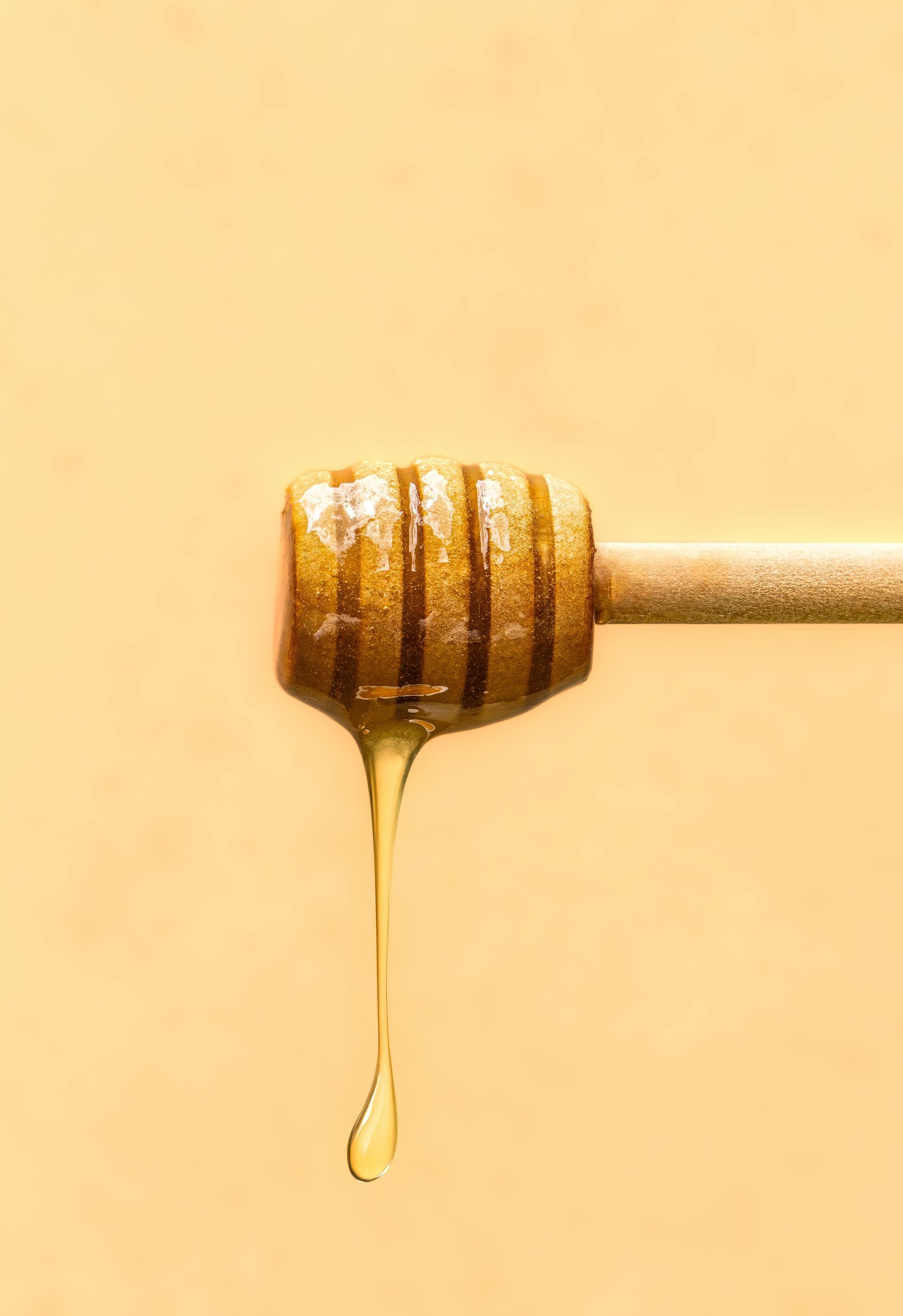 A wooden honey dipper drizzling golden honey against a light yellow background.