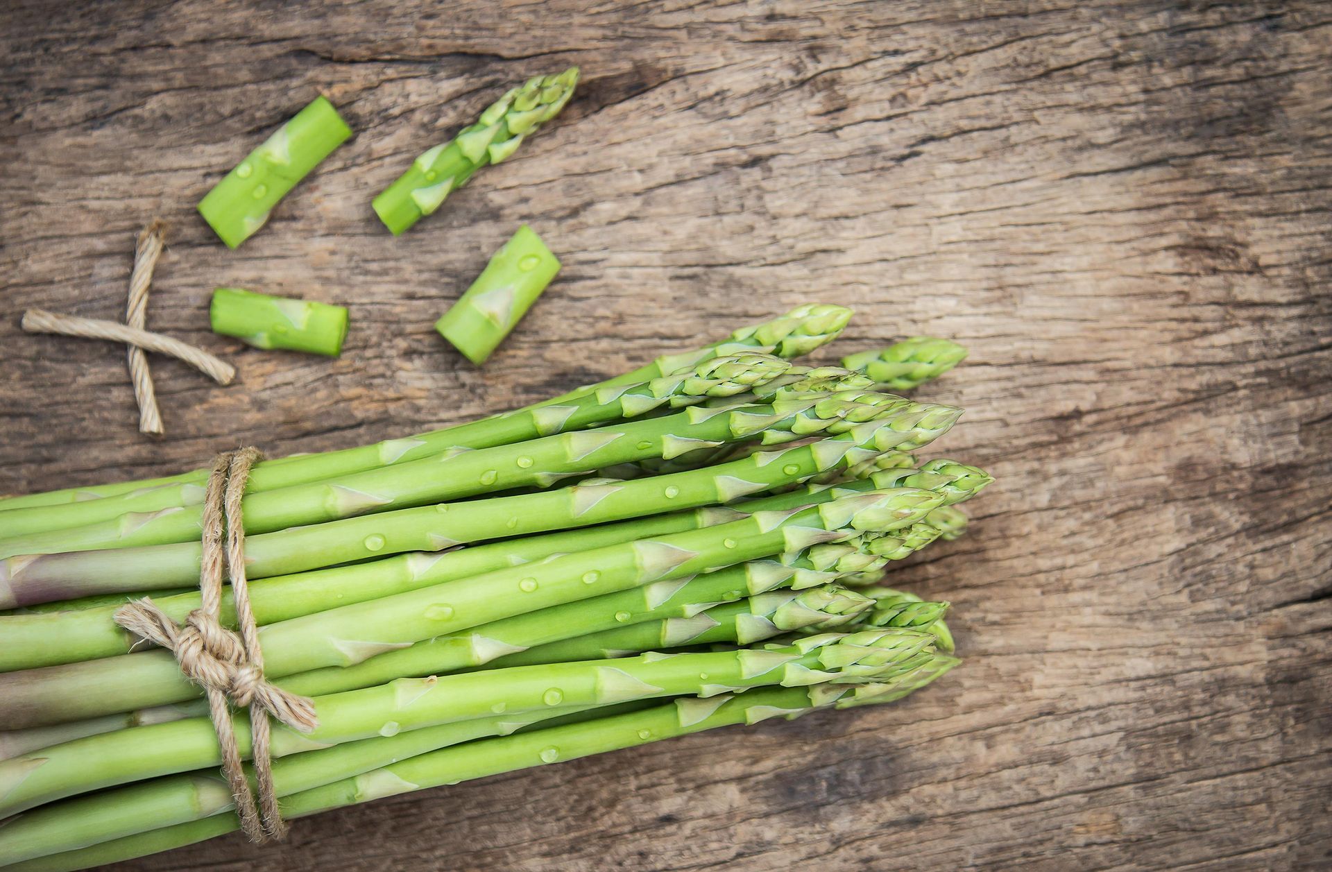 A bundle of fresh green asparagus tied with twine on a rustic wooden background, with several cut pieces nearby.