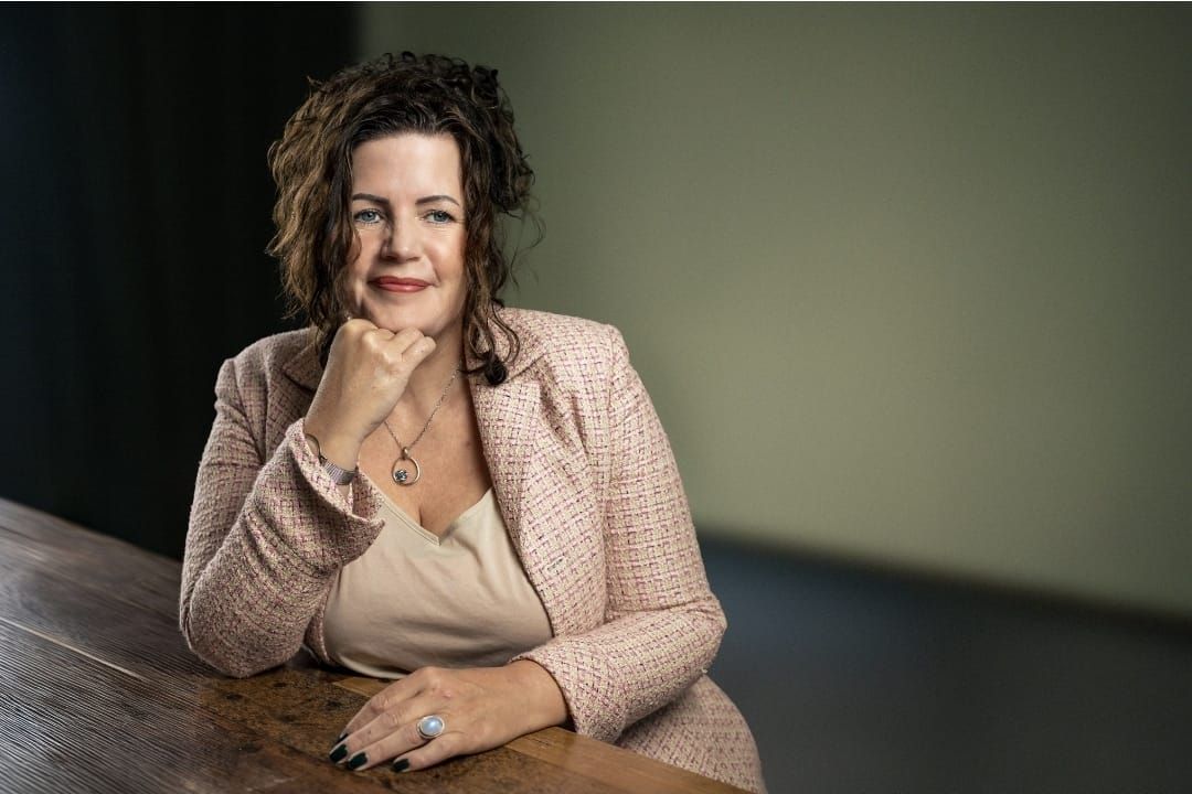 Een vrouw in een roze trui rust met haar kin op haar hand en staart peinzend in een zacht verlichte kamer.