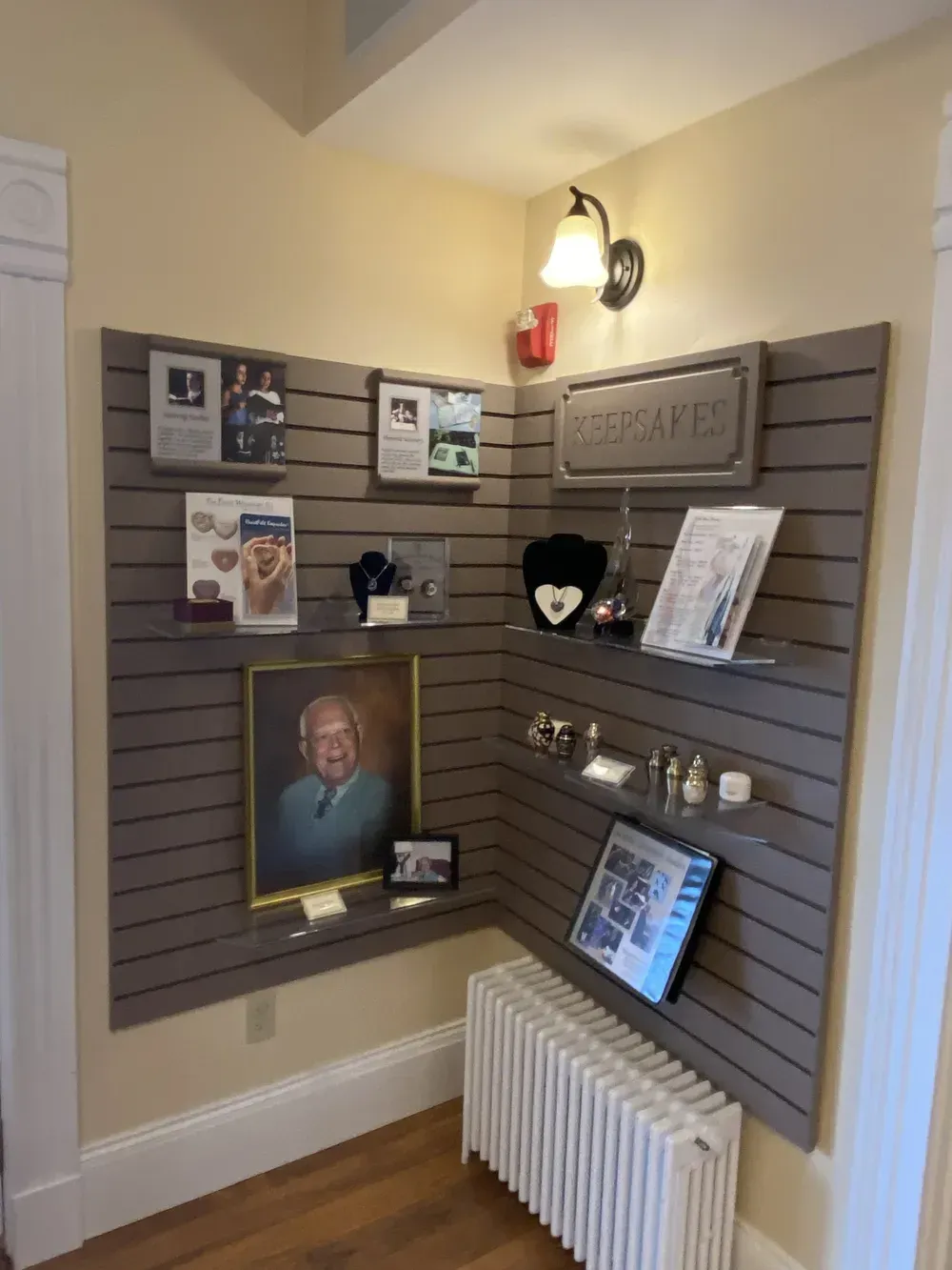 Memorial corner with framed photos, cards, flowers, and a radiator in a beige hallway corner