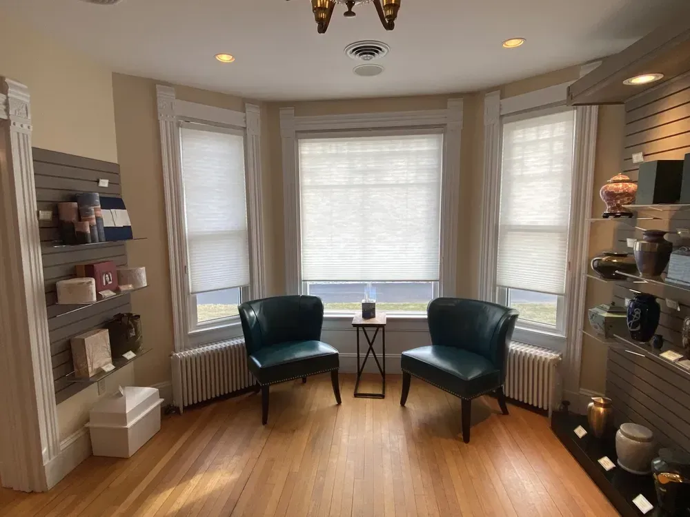 Cozy bay-window sitting room with two teal chairs, small table, built-in shelves, and warm wood flooring.