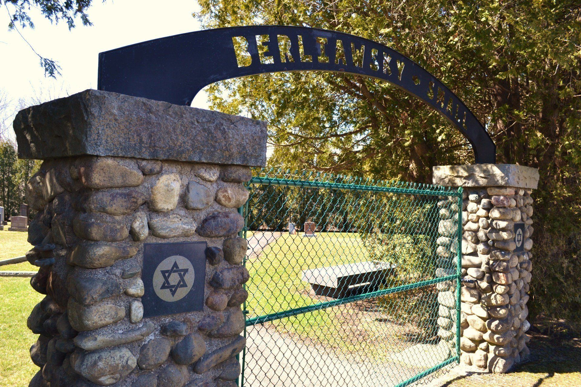 Stone entrance gate with metal arch and green fence, leading to a park path and picnic table