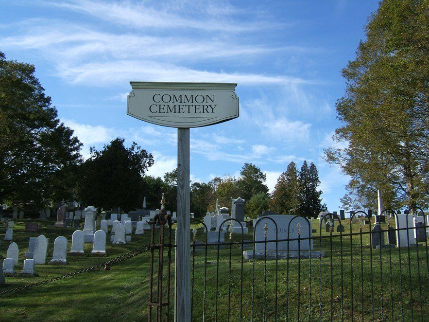 Entrance sign to Common Ground Cemetery with rows of gravestones and trees under a blue sky