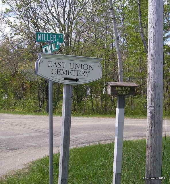 Street signs for Miller Rd and East Union Cemetery beside a road with trees and grass.