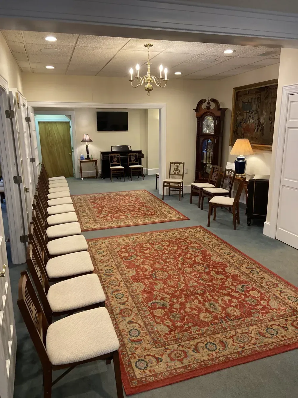 Long room with patterned rugs, row of chairs, desk, chandelier, and wall-mounted TV.