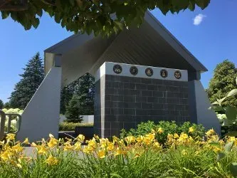 Memorial monument with black stone wall and circular plaques, framed by yellow flowers and trees under a blue sky