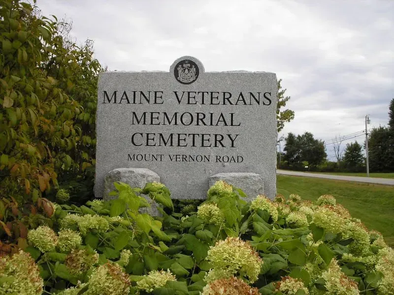 Stone sign reading “Maine Veterans Memorial Cemetery” beside green shrubs and a roadside
