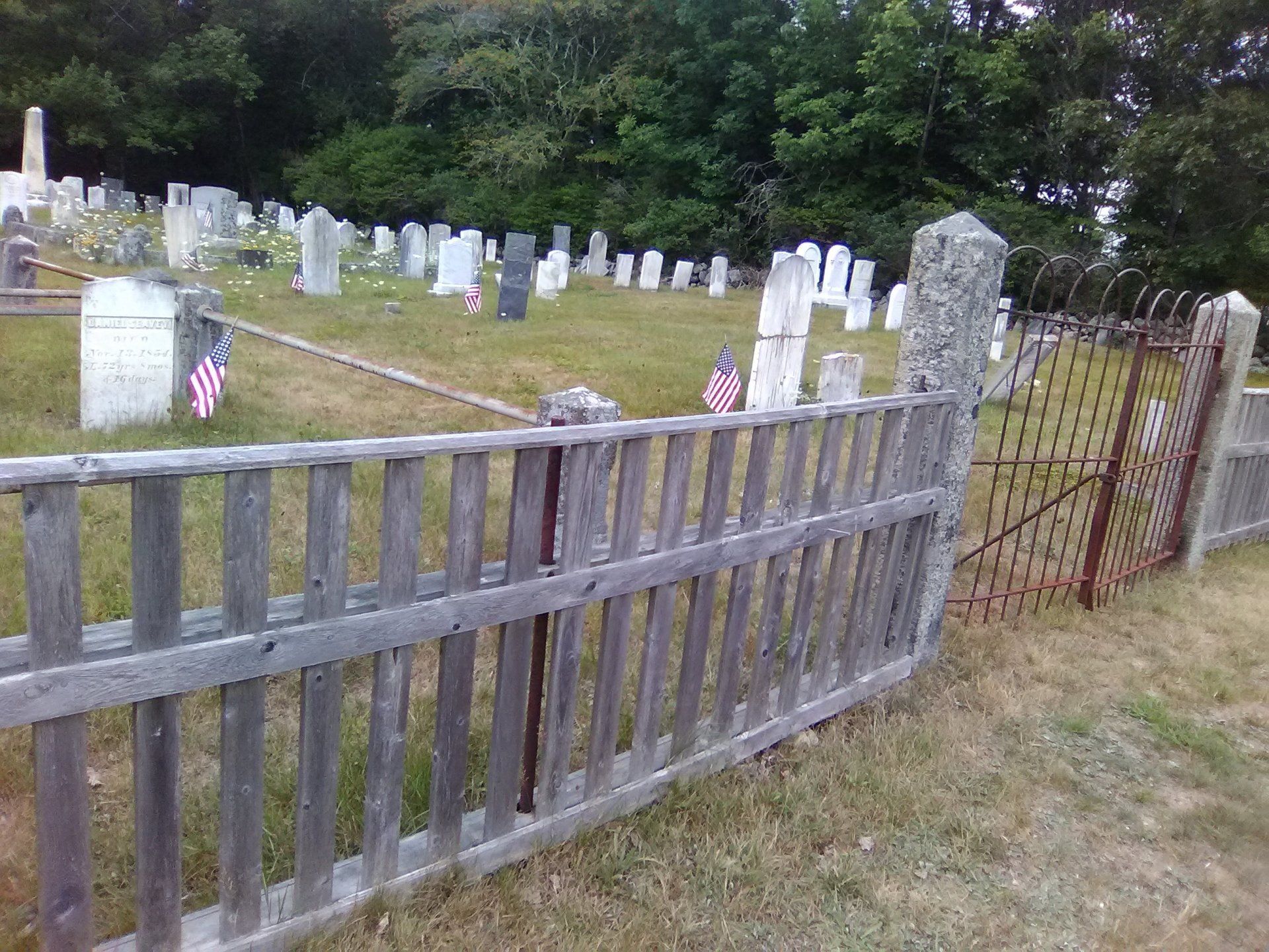 Wooden fence beside a small graveyard with white headstones and American flags, bordered by trees.