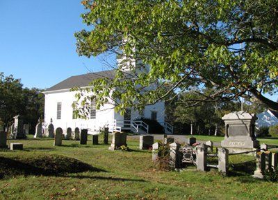 White church with graveyard and tombstones under a large tree on a sunny day