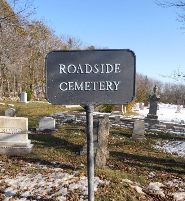 Roadside Cemetery sign in a small graveyard with headstones and bare trees under a blue sky.