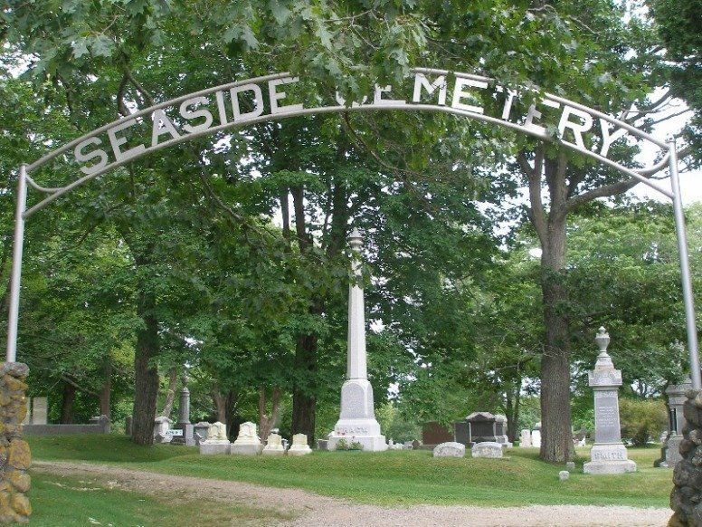Entrance arch reading “Seaside Cemetery” over a tree-lined cemetery with gravestones and a monument