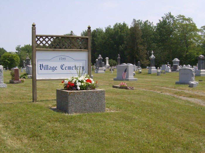 Village cemetery sign beside a flower planter, with gravestones scattered across a grassy field