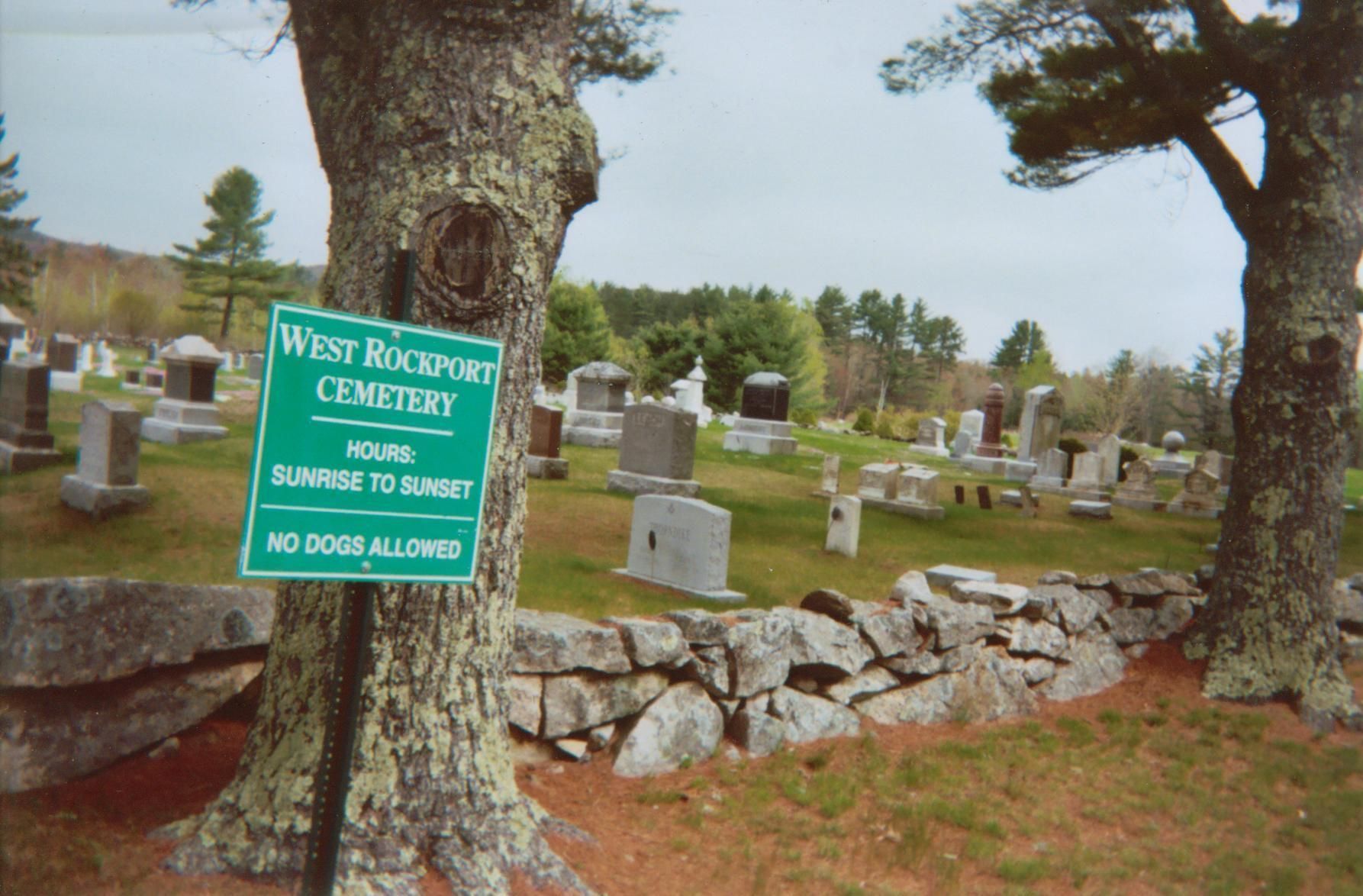 West Rockland Cemetery sign beside a stone wall among tombstones in a grassy graveyard.
