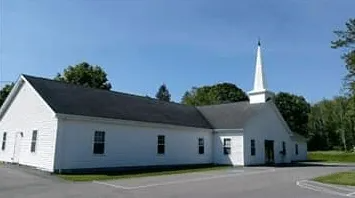 White church with tall steeple beside a parking lot, surrounded by trees under a clear blue sky