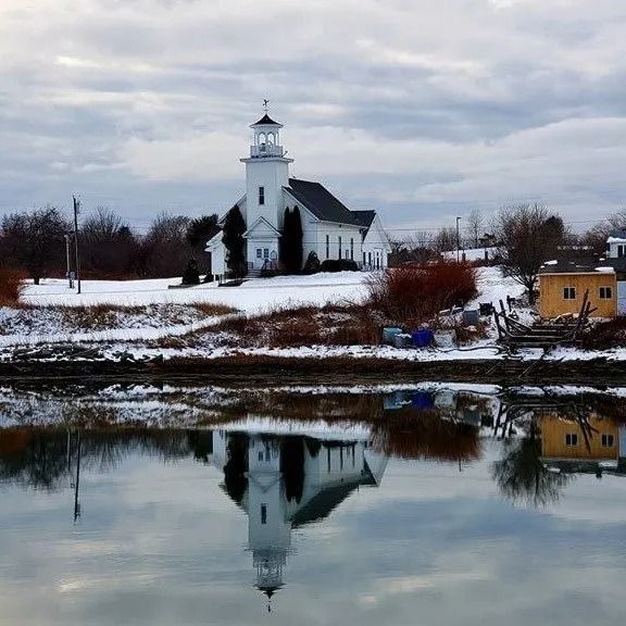 White church on snowy shore, reflected in calm water under a cloudy winter sky