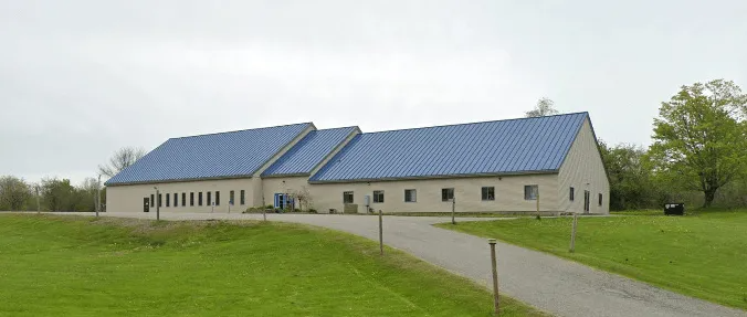 Long beige building with a blue roof beside a gravel road and green lawn under cloudy skies