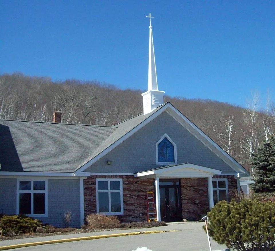 Brick church with a white steeple and blue-gray siding in front of a wooded hillside