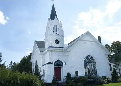 White church with a tall steeple and red door against a blue sky, surrounded by greenery