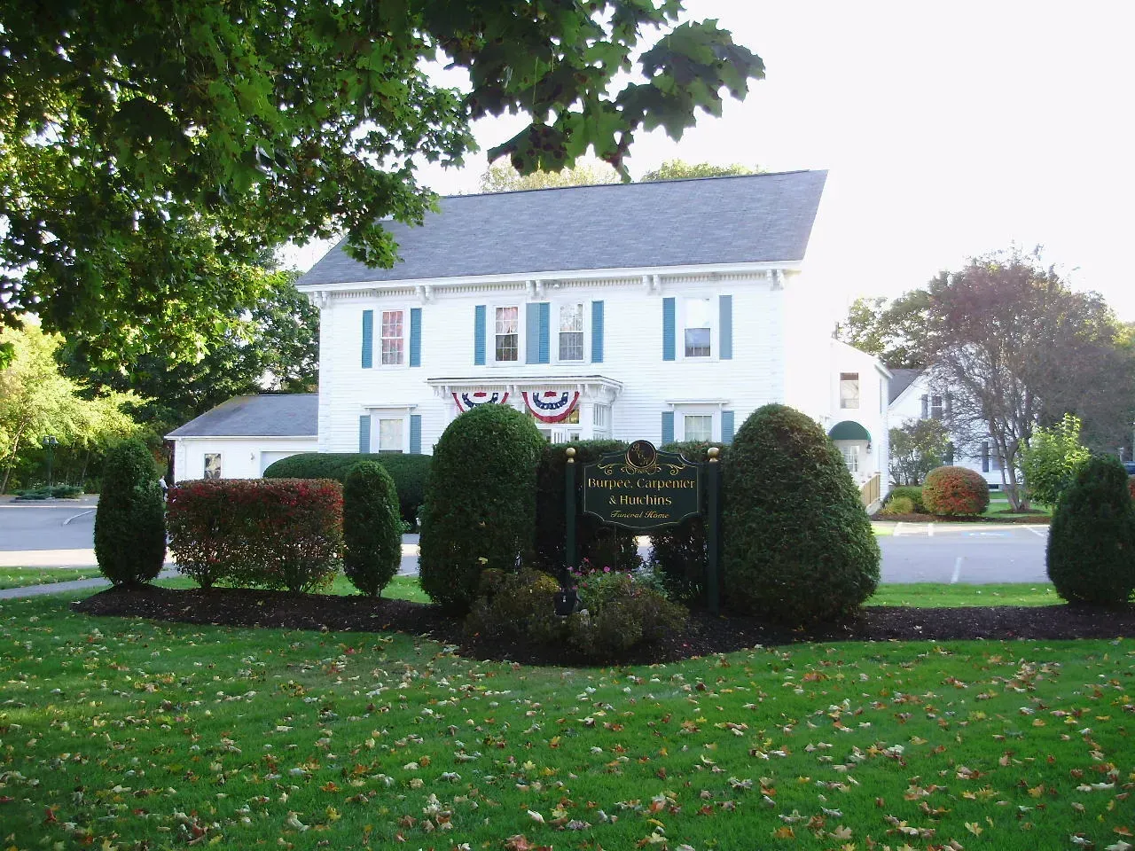 White house with blue shutters behind trimmed hedges and a green lawn under trees
