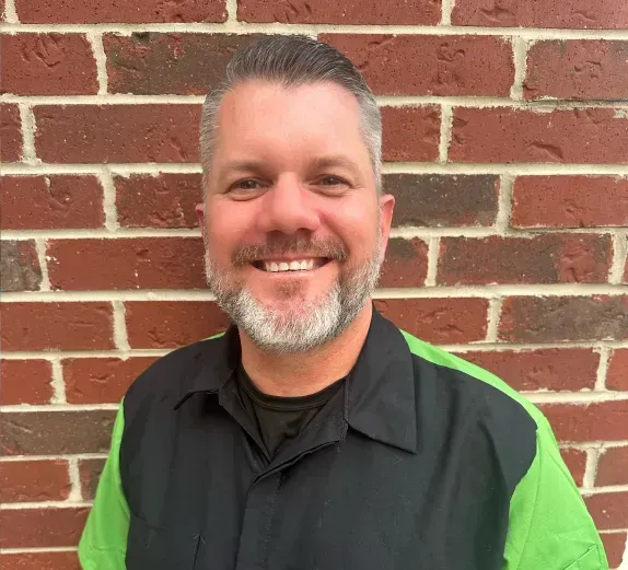 Smiling person in a black shirt with neon green sleeves standing in front of a brick wall | Perryman Auto Repair