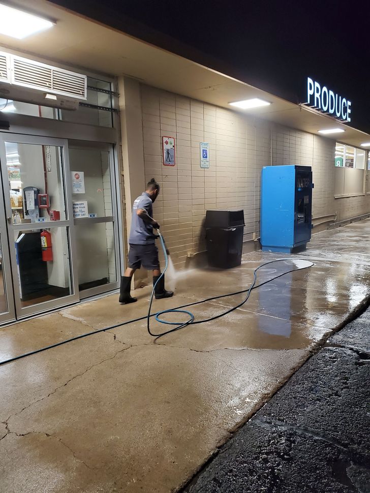 Person spraying down a wet storefront walkway beside blue recycling bins at night