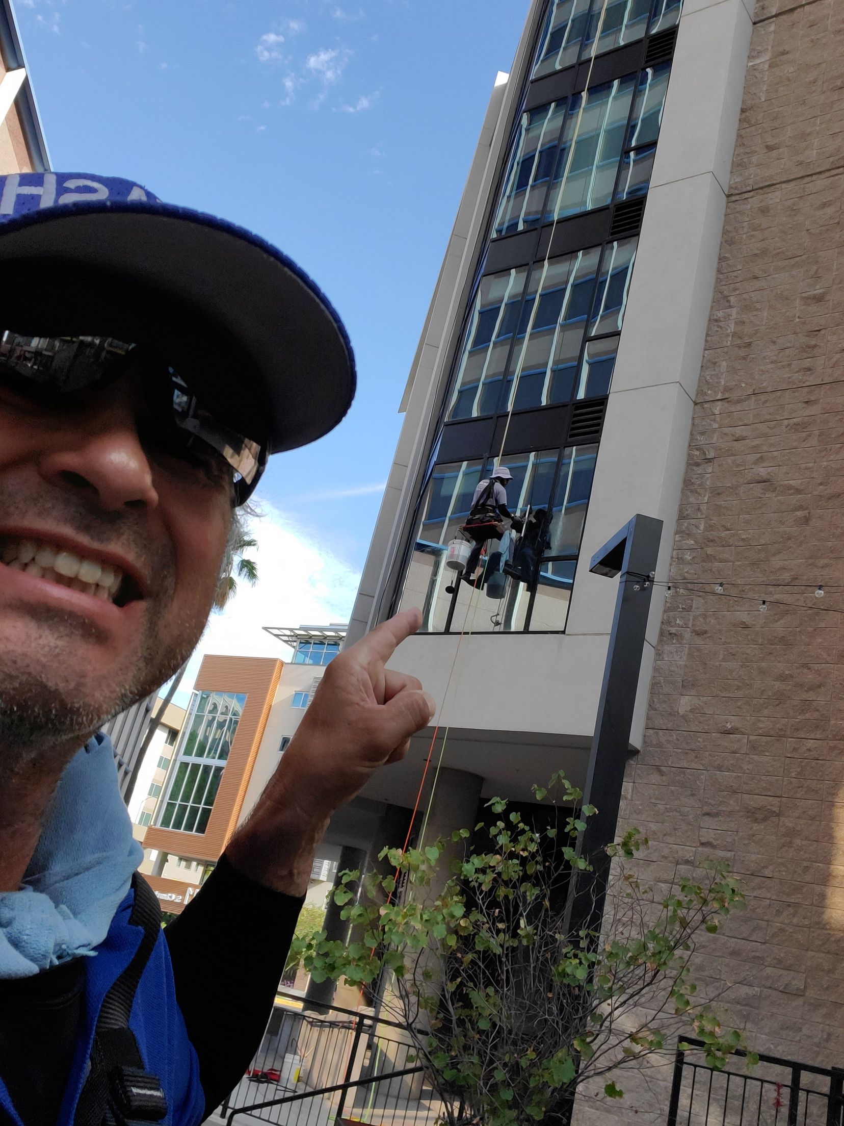 Man in blue cap and sunglasses pointing at a tall glass building with people on a balcony.