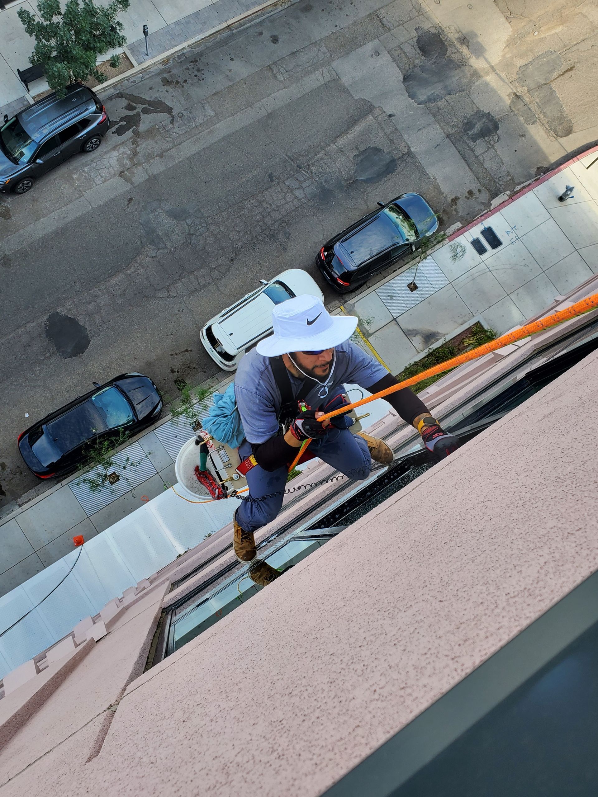Worker on a rope access harness cleaning a building window from above a street with parked cars.