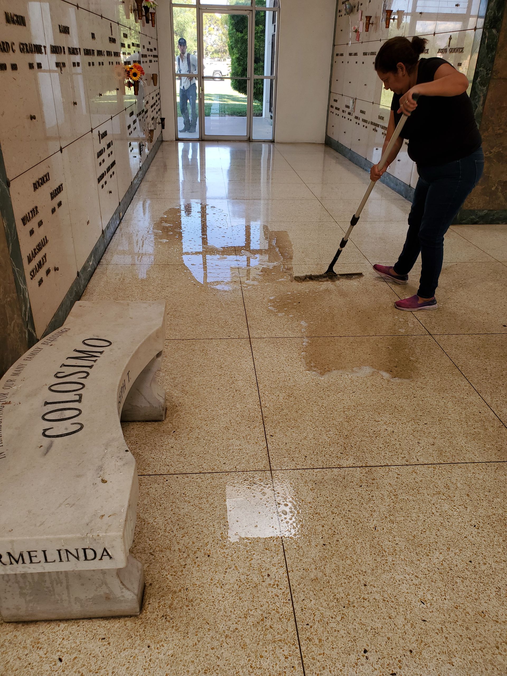 Person mopping a wet hallway floor beside a bench in a bright indoor corridor.