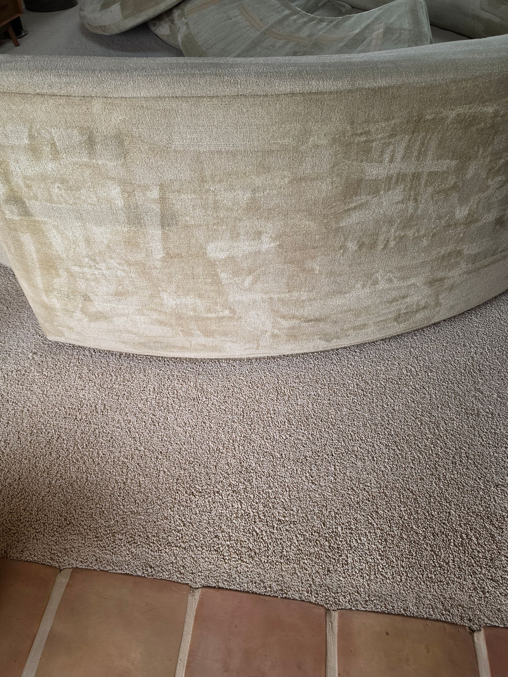 Close-up of a beige upholstered couch above a speckled carpet and terracotta tile floor.