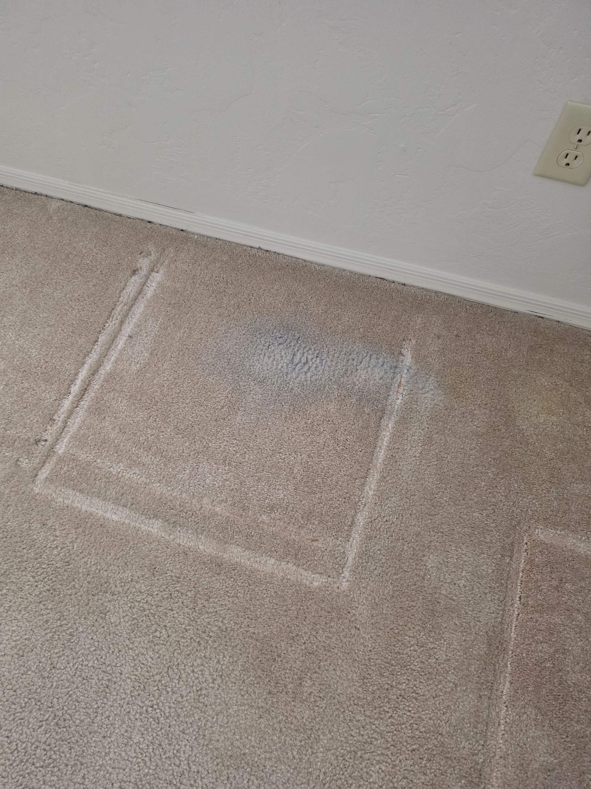 Carpeted floor corner with white chalk lines marking a rectangle near a wall outlet.