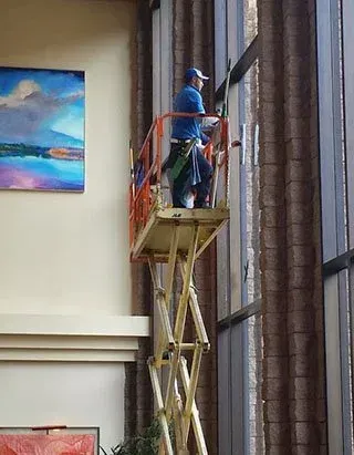 Worker on a lift cleaning tall indoor windows beside a wall painting