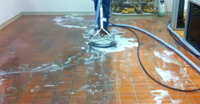 Person pressure-washing a driveway beside a metal fence, wearing yellow gloves and boots.
