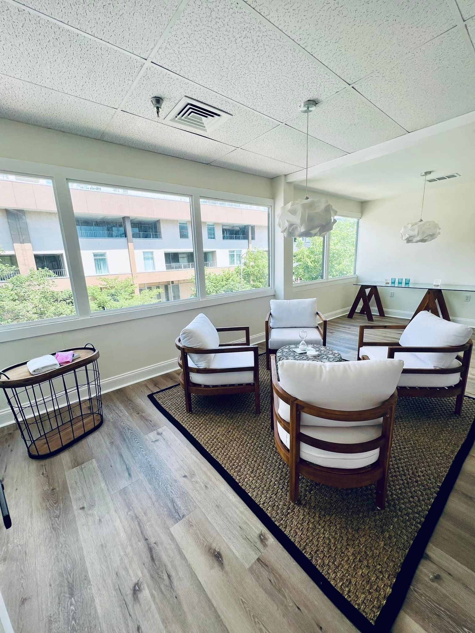 A sunlit room with four white chairs arranged around a round rug on a wood floor, with large windows overlooking trees.