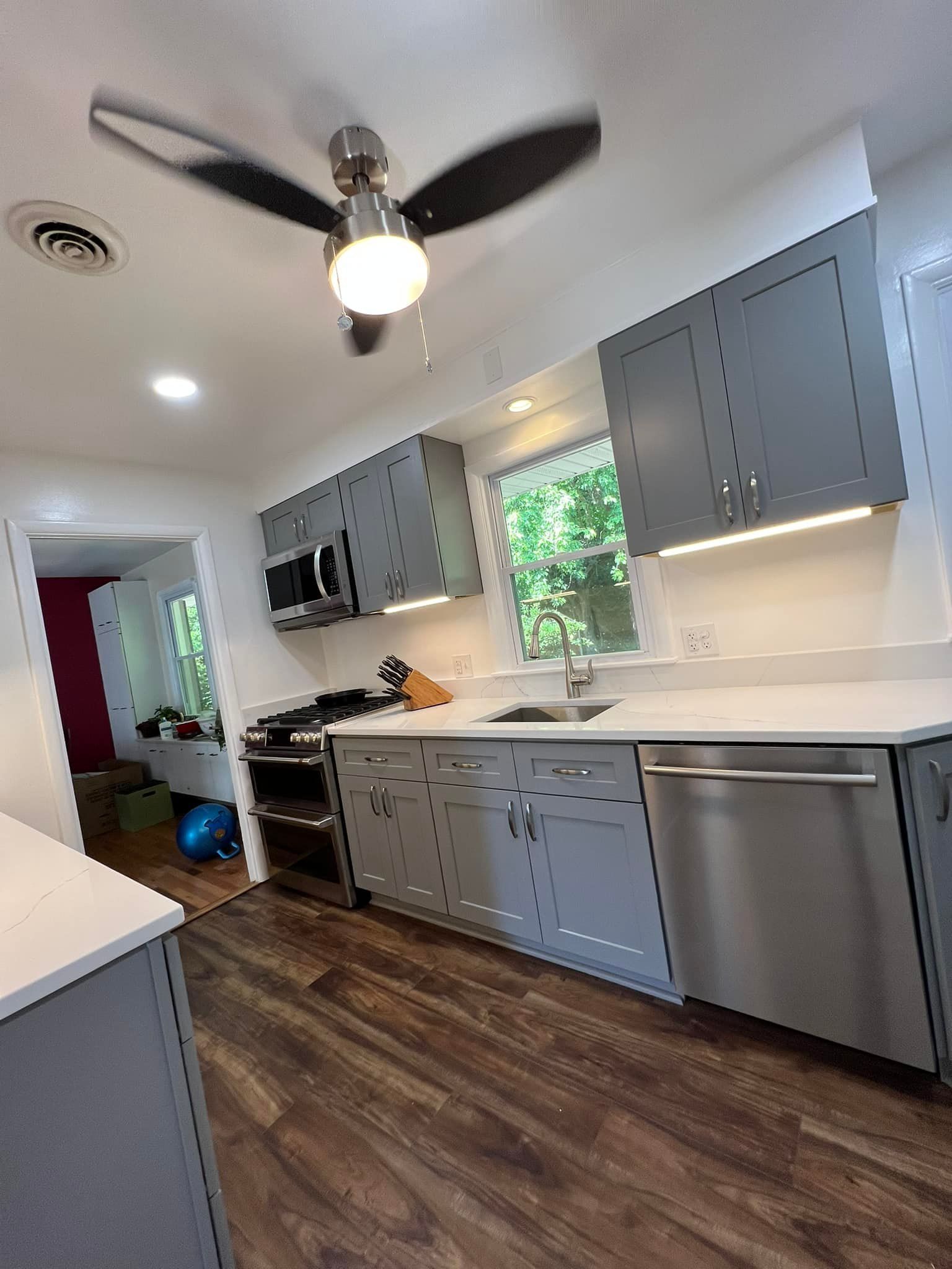A modern kitchen featuring grey cabinets, a stainless steel dishwasher, and a dark ceiling fan over wood-look flooring.