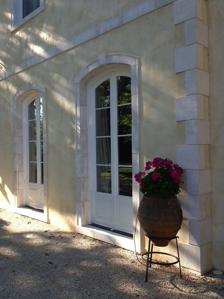 Two white arched French doors set in a beige wall with stone corner trim, featuring a potted plant on a metal stand.