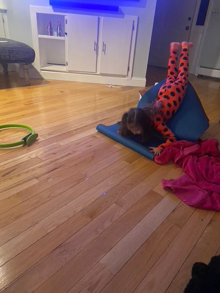 A child in spotted pajamas performs a handstand on a blue mat in a living room with wooden floors.