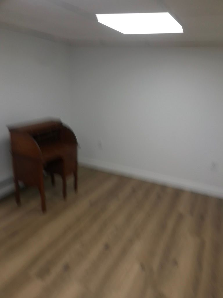 A dark wooden roll-top desk sits in the corner of a sparsely furnished room with light wood flooring and white walls.