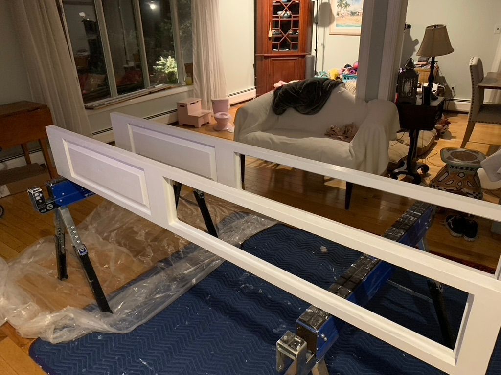 A white cabinet door panel rests on blue sawhorses in a living room, set up for painting on a protective drop cloth.