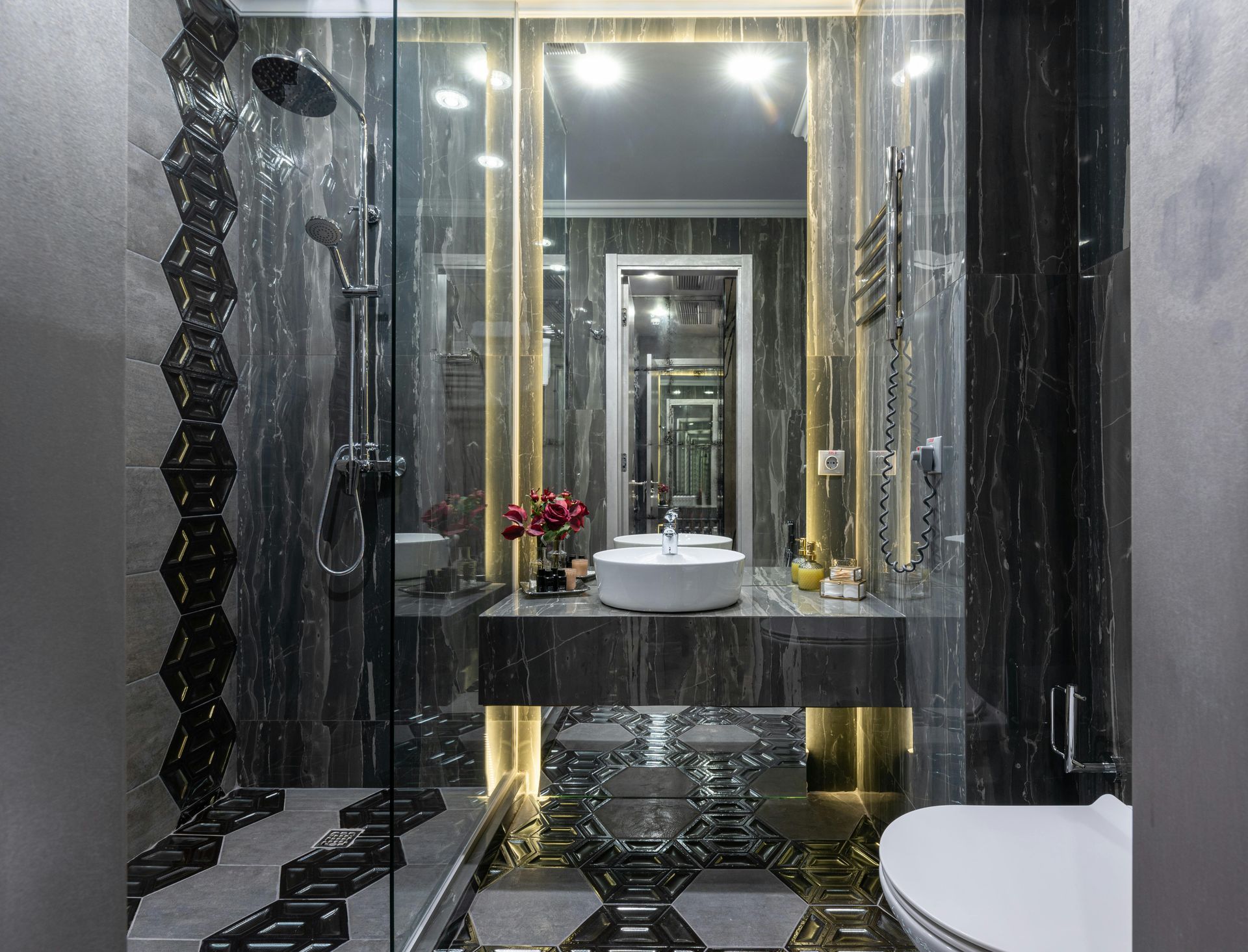 A modern luxury bathroom featuring dark marble walls, a glass-enclosed shower, a white vessel sink, and gold accents.