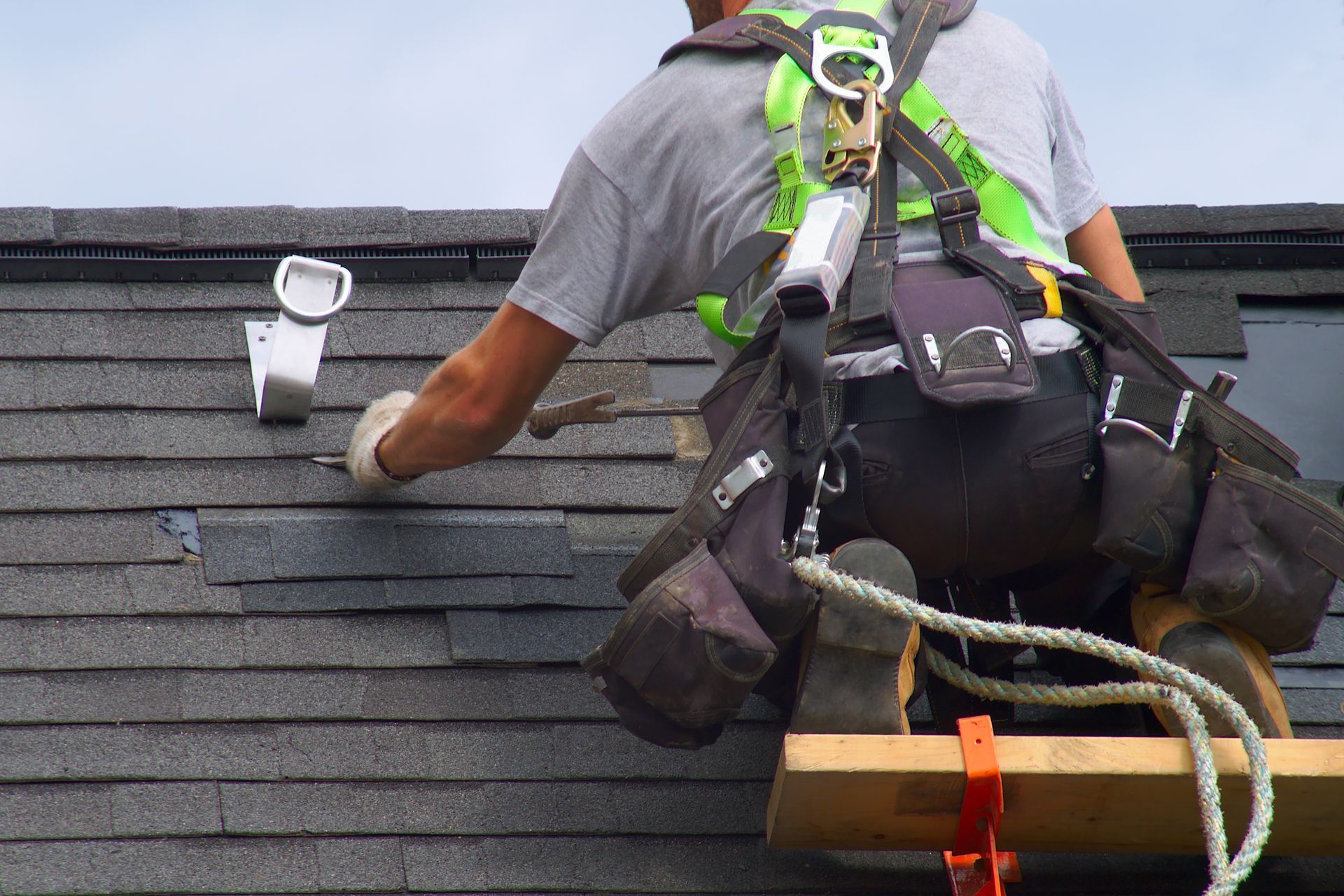 construction worker installing asphalt shingles on a roof while wearing a safety harness.