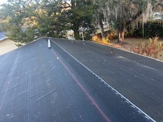 Black roofing material on a house roof. Trees and sky in the background.