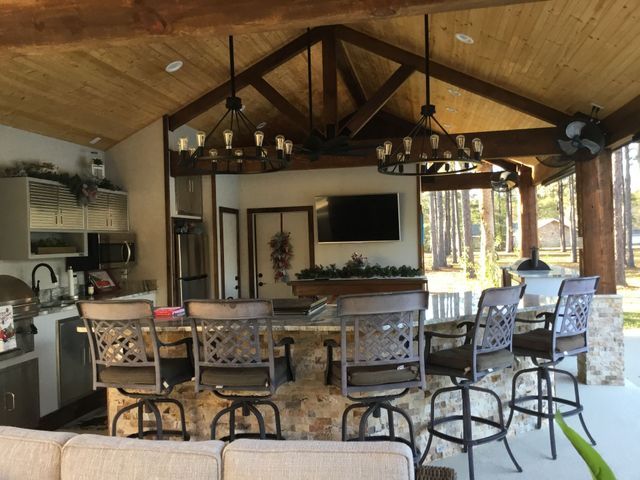 Covered outdoor kitchen and bar with stools, ceiling fans, and a TV under a wooden pavilion.