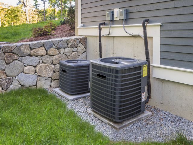 Two outdoor air conditioning units beside a house and stone retaining wall