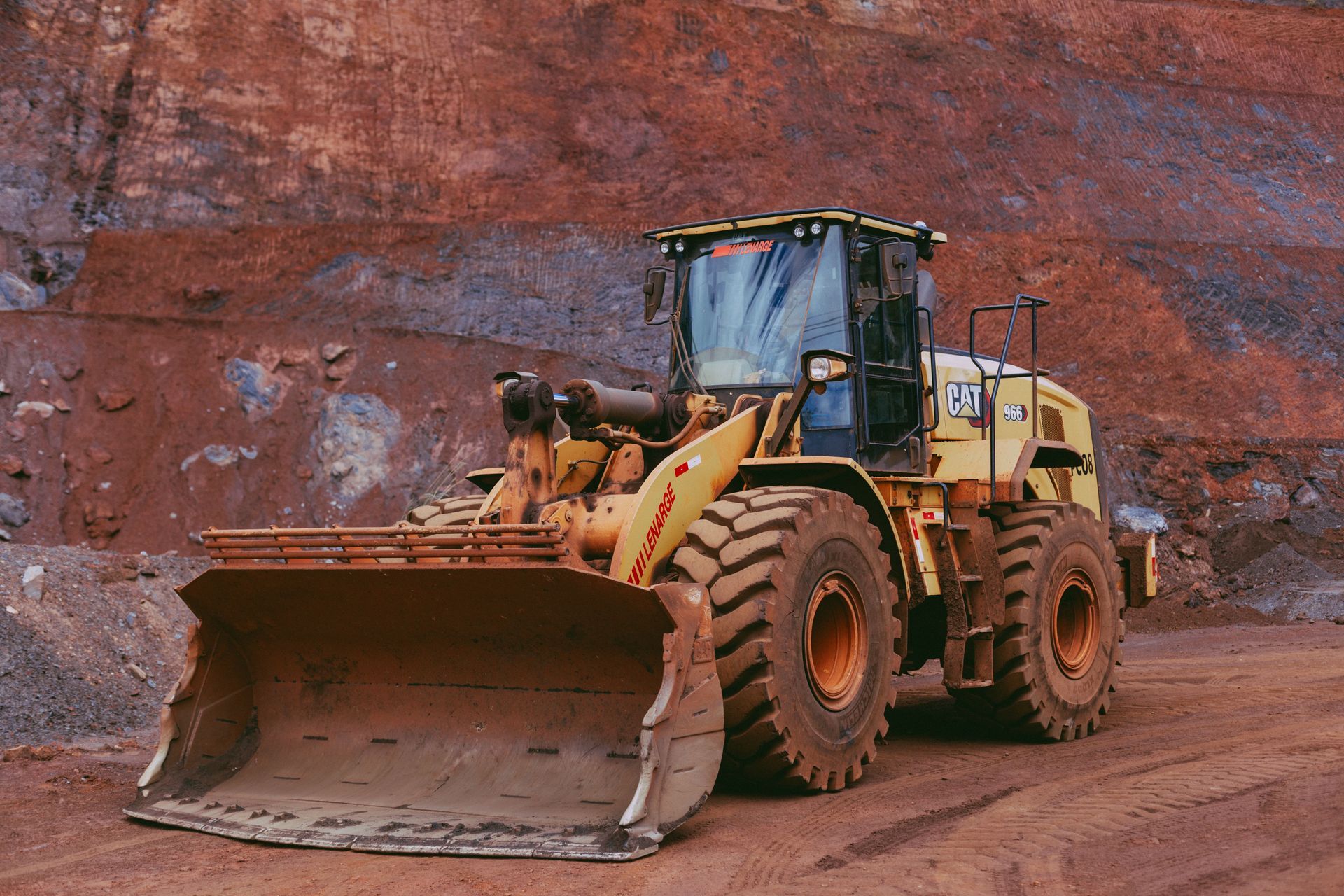 Yellow front-end loader in a rocky quarry, with a large bucket on reddish dirt.