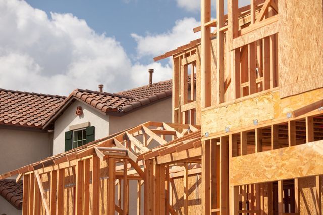 Wood-framed house under construction beside a finished home, with a red-tile roof and blue sky.